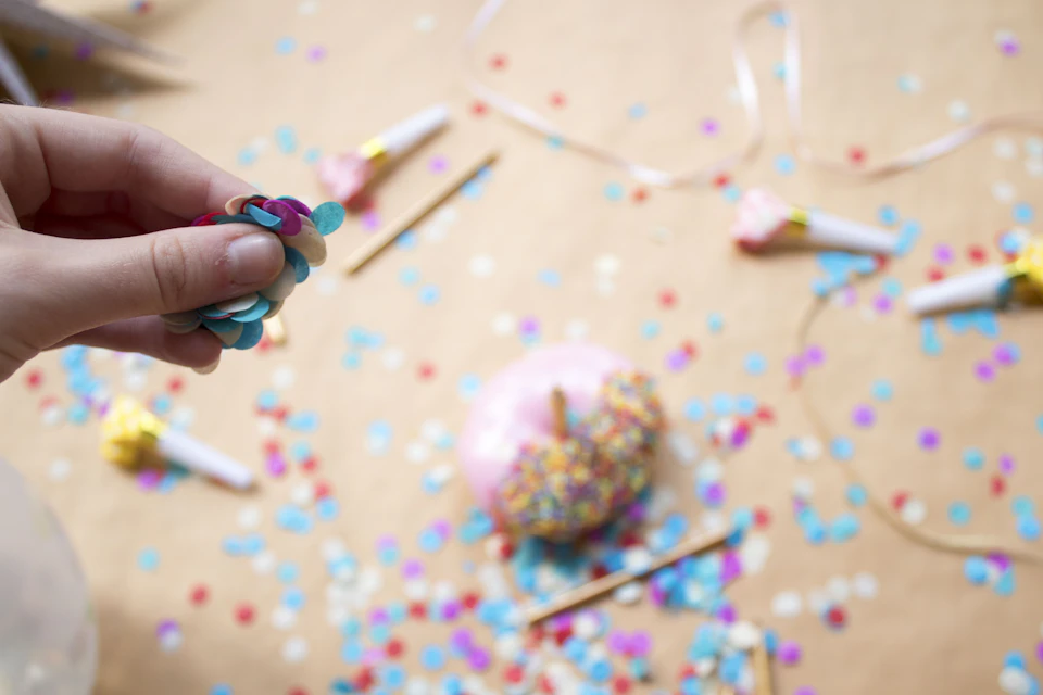Hand holding colorful party decoration made of confetti and a pink donut amidst scattered confetti