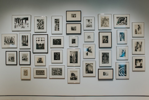 A gallery wall of various framed black and white photographs and artworks arranged in an irregular grid, showcasing a variety of styles and subjects. The frames are mostly simple and understated, allowing the artworks to be the central focus. The wall is painted a neutral color, enhancing the contrast of the monochrome images.