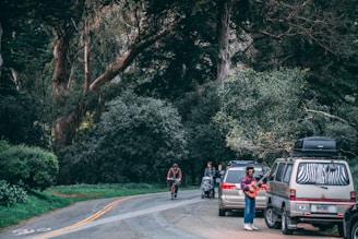 two vehicle park beside the road with people passing by surrounded with trees