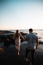 A couple is walking barefoot on rocky terrain by the ocean, holding hands. The sun is setting, creating a warm glow on the surroundings. The woman's long hair flows down her back, and they appear to be enjoying a serene moment together.