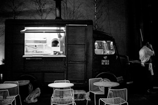 A vintage-style food truck is parked in a dimly lit area with an open window revealing the kitchen inside. There are metal chairs and round tables set in front of the truck, suggesting an outdoor dining area. A person in a white shirt stands beside the truck, possibly a vendor or customer, engaged in a phone conversation.