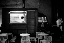 A vintage-style food truck is parked in a dimly lit area with an open window revealing the kitchen inside. There are metal chairs and round tables set in front of the truck, suggesting an outdoor dining area. A person in a white shirt stands beside the truck, possibly a vendor or customer, engaged in a phone conversation.