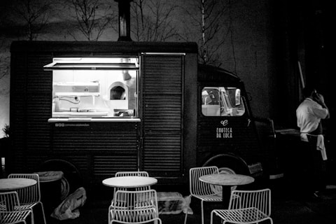 A vintage-style food truck is parked in a dimly lit area with an open window revealing the kitchen inside. There are metal chairs and round tables set in front of the truck, suggesting an outdoor dining area. A person in a white shirt stands beside the truck, possibly a vendor or customer, engaged in a phone conversation.