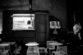 A vintage-style food truck is parked in a dimly lit area with an open window revealing the kitchen inside. There are metal chairs and round tables set in front of the truck, suggesting an outdoor dining area. A person in a white shirt stands beside the truck, possibly a vendor or customer, engaged in a phone conversation.