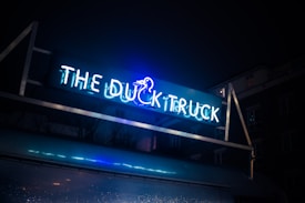 A neon sign displayed at night with the words 'THE DUCK TRUCK' in bright blue and white colors. The sign includes an illustration of a duck, and the structure is set against a dark sky with visible building silhouettes in the background.