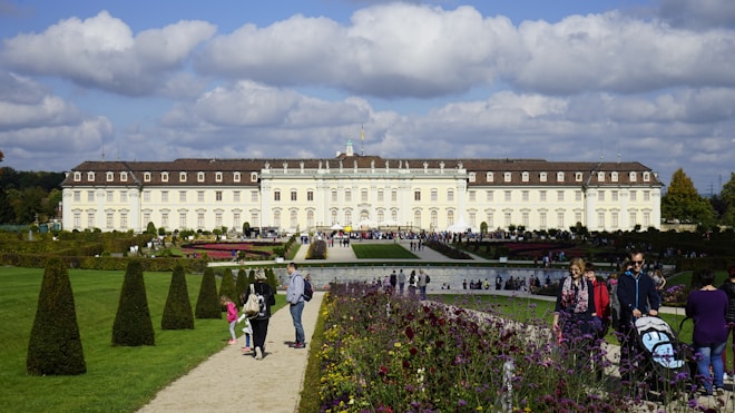 A large, ornate palace with a symmetrical design and numerous windows stretches across the center of the image. In front, expansive gardens feature neatly trimmed hedges, colorful flowers, and a wide pathway lined with people walking and enjoying the scenery. The sky is dotted with fluffy white clouds.