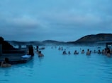People relaxing in a large geothermal spa surrounded by steam, with views of distant hills under a cloudy sky. The atmosphere is serene and the blue water is inviting.