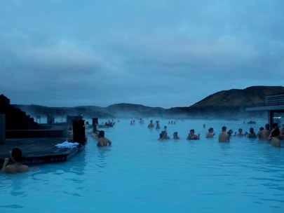 People relaxing in a large geothermal spa surrounded by steam, with views of distant hills under a cloudy sky. The atmosphere is serene and the blue water is inviting.