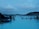 People relaxing in a large geothermal spa surrounded by steam, with views of distant hills under a cloudy sky. The atmosphere is serene and the blue water is inviting.