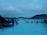 People relaxing in a large geothermal spa surrounded by steam, with views of distant hills under a cloudy sky. The atmosphere is serene and the blue water is inviting.