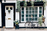A charming exterior featuring a white door with a brass mail slot and handle, adorned with a basket of flowers. Next to the door, a chalkboard sign reads 'No. 4 Holywell B&B.' Beside it, a large window with multiple panes has hanging plants and floral decorations above. A vintage bicycle is parked with flowers in its basket, adding to the quaint, inviting ambiance.