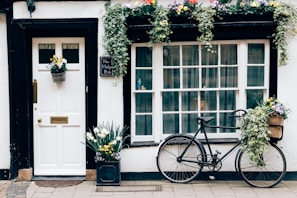 A charming exterior featuring a white door with a brass mail slot and handle, adorned with a basket of flowers. Next to the door, a chalkboard sign reads 'No. 4 Holywell B&B.' Beside it, a large window with multiple panes has hanging plants and floral decorations above. A vintage bicycle is parked with flowers in its basket, adding to the quaint, inviting ambiance.