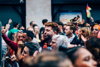 A diverse group of people gathered closely together, suggesting a lively outdoor event or festival. Faces show expressions of joy and engagement, with some individuals adorned with face paint. Bright colors are visible, including a small rainbow flag waving in the background.