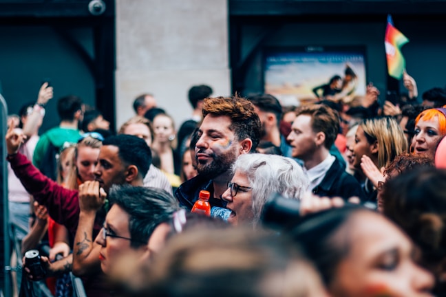 A diverse group of people gathered closely together, suggesting a lively outdoor event or festival. Faces show expressions of joy and engagement, with some individuals adorned with face paint. Bright colors are visible, including a small rainbow flag waving in the background.