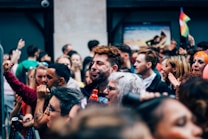 A diverse group of people gathered closely together, suggesting a lively outdoor event or festival. Faces show expressions of joy and engagement, with some individuals adorned with face paint. Bright colors are visible, including a small rainbow flag waving in the background.