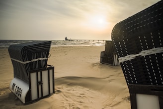 Wicker beach chairs are positioned on a sandy shore, with a calm sea and a pier stretching out in the background. The sun is setting or rising, casting a warm, golden hue over the scene.