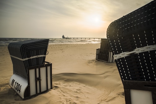 Wicker beach chairs are positioned on a sandy shore, with a calm sea and a pier stretching out in the background. The sun is setting or rising, casting a warm, golden hue over the scene.