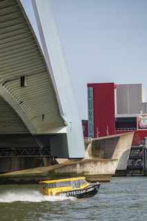 A modern bridge with a large, angled beam dominates the left side of the image. Below, a water taxi with a bright yellow roof and dark hull speeds along the river, creating a frothy wake. In the background, a red building with the letters LUXOR and additional modern architectural elements are visible.
