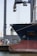 A large ship is docked at a port, with its hull prominently visible. In the background, there is an industrial crane used for loading and unloading cargo. The scene suggests a busy harbor setup with various equipment and structures surrounding the ship.
