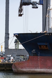 A large ship is docked at a port, with its hull prominently visible. In the background, there is an industrial crane used for loading and unloading cargo. The scene suggests a busy harbor setup with various equipment and structures surrounding the ship.