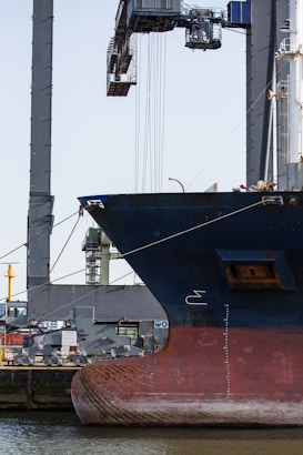 A large ship is docked at a port, with its hull prominently visible. In the background, there is an industrial crane used for loading and unloading cargo. The scene suggests a busy harbor setup with various equipment and structures surrounding the ship.