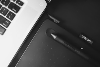 A portion of a laptop keyboard is visible on the left with a focus on keys such as delete, enter, and return. Adjacent to the keyboard, a black drawing tablet and stylus pen are present, with the stylus resting on the tablet. The branding 'Wacom' is visible on a USB drive next to the tablet.