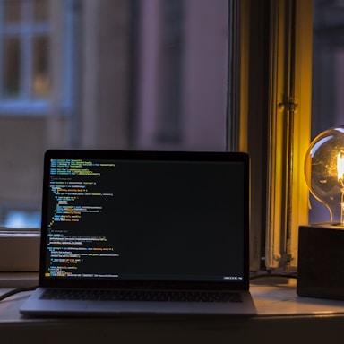 A dimly lit room with a laptop on a window sill displaying lines of code on the screen. Beside the laptop, an exposed incandescent light bulb glows warmly, casting soft light and shadows.