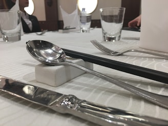 A dining table is elegantly set with silverware including a knife, fork, and spoon. The spoon rests on a small rectangular block. There are clear drinking glasses placed around the table, with some folded napkins visible in the background. The tablecloth has a subtle wave pattern. Part of a person is visible, dressed in formal attire.