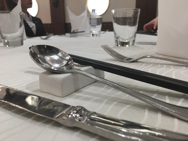 A dining table is elegantly set with silverware including a knife, fork, and spoon. The spoon rests on a small rectangular block. There are clear drinking glasses placed around the table, with some folded napkins visible in the background. The tablecloth has a subtle wave pattern. Part of a person is visible, dressed in formal attire.