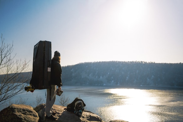 A person stands atop a rocky outcrop overlooking a serene lake with a forested hill in the background. The person is wearing outdoor gear, including a backpack and a thick coat, and seems to be carrying a large, rectangular item on their back. The sun is shining brightly, reflecting off the lake's surface, creating a glistening effect.