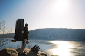 A person stands atop a rocky outcrop overlooking a serene lake with a forested hill in the background. The person is wearing outdoor gear, including a backpack and a thick coat, and seems to be carrying a large, rectangular item on their back. The sun is shining brightly, reflecting off the lake's surface, creating a glistening effect.