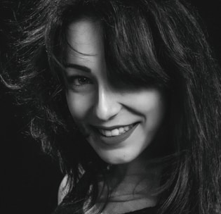 A black and white portrait of a person with long, dark hair and a bright smile. The hair is voluminous and slightly tousled, partially covering one eye. The lighting creates contrast, highlighting the features and hair texture against a dark background.