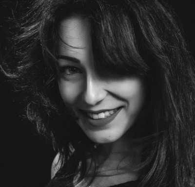 A black and white portrait of a person with long, dark hair and a bright smile. The hair is voluminous and slightly tousled, partially covering one eye. The lighting creates contrast, highlighting the features and hair texture against a dark background.