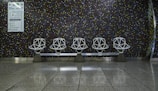 Five geometric white chairs are arranged in a row against a dark, tiled wall with a mosaic pattern of small colored squares. The floor is polished and reflective, adding to the modern aesthetic. A sign with directions is visible on the left side of the wall, indicating a public space like a waiting area or transportation terminal.