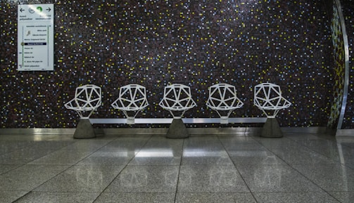Five geometric white chairs are arranged in a row against a dark, tiled wall with a mosaic pattern of small colored squares. The floor is polished and reflective, adding to the modern aesthetic. A sign with directions is visible on the left side of the wall, indicating a public space like a waiting area or transportation terminal.