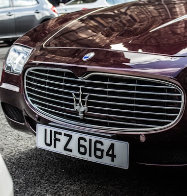 A close-up of the front grille of a luxury car with a distinctive trident logo, reflecting the cityscape. The vehicle's license plate reads UFZ 6164, and the body is a deep burgundy color.