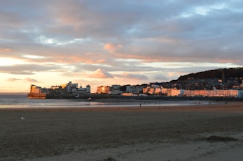 A coastal town is bathed in the warm colors of sunset, with a calm sea and sandy beach in the foreground. The sky is scattered with fluffy clouds reflecting the orange and pink hues of the setting sun. Buildings line the waterfront, illuminated by the golden light, creating a serene and picturesque scene. Sparse figures can be seen walking along the shore.