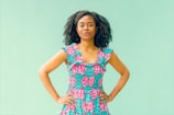 A person stands confidently with hands on hips, wearing a colorful, patterned dress with a vibrant pink and teal design. The background is a solid mint green color.