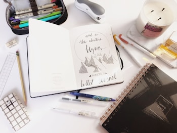 An open notebook with creative lettering displaying 'the adventure begins 2018' and mountain illustrations is surrounded by various stationery items. There is a pencil case filled with markers, a white candle, some pens, and a closed black spiral-bound planner. Additional items include a gold perfume bottle, a white ruler, and a small grid-patterned notepad.