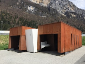 Two modern, rust-colored structures with a minimalist design are situated near a rocky mountain landscape. The buildings have rectangular shapes with small vertical openings. The surrounding area includes green grass and trees against a rocky cliff backdrop, under a partly cloudy sky.