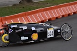 A streamlined, low-profile vehicle with the number 524 displayed on the side is moving along a paved road. The vehicle has a black body with various sponsor logos on it, including a prominent Shell logo. It features two large, spoked wheels at the back and one small wheel at the front, indicating it is designed for efficiency, possibly a prototype or eco-marathon vehicle. In the background, there are orange barriers lining the edge of the track, and a grassy area can be seen.