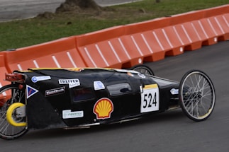 A streamlined, low-profile vehicle with the number 524 displayed on the side is moving along a paved road. The vehicle has a black body with various sponsor logos on it, including a prominent Shell logo. It features two large, spoked wheels at the back and one small wheel at the front, indicating it is designed for efficiency, possibly a prototype or eco-marathon vehicle. In the background, there are orange barriers lining the edge of the track, and a grassy area can be seen.