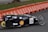 A streamlined, low-profile vehicle with the number 524 displayed on the side is moving along a paved road. The vehicle has a black body with various sponsor logos on it, including a prominent Shell logo. It features two large, spoked wheels at the back and one small wheel at the front, indicating it is designed for efficiency, possibly a prototype or eco-marathon vehicle. In the background, there are orange barriers lining the edge of the track, and a grassy area can be seen.
