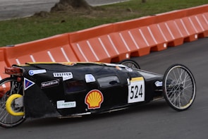 A streamlined, low-profile vehicle with the number 524 displayed on the side is moving along a paved road. The vehicle has a black body with various sponsor logos on it, including a prominent Shell logo. It features two large, spoked wheels at the back and one small wheel at the front, indicating it is designed for efficiency, possibly a prototype or eco-marathon vehicle. In the background, there are orange barriers lining the edge of the track, and a grassy area can be seen.