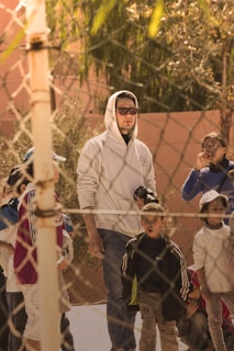 A group of children are gathered outdoors on a sunny day, with a man in a white hoodie and sunglasses standing among them. A chain-link fence is in the foreground, and trees can be seen in the background, casting shadows on the scene.