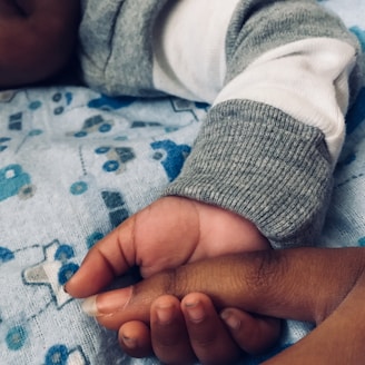 A close-up view of an infant's hand resting on an adult's hand. The infant is wearing a gray and white sleeve, and the background features a gray fabric with blue toy vehicle patterns.