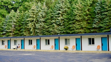 A single-story motel building with a row of five doors and windows, each featuring bright blue doors and orange chairs placed outside. White potted flowers are seen beside a person seated near one of the rooms. The background is dominated by a dense line of tall evergreen trees, adding a natural feel to the setting.