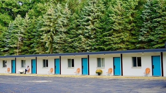 A single-story motel building with a row of five doors and windows, each featuring bright blue doors and orange chairs placed outside. White potted flowers are seen beside a person seated near one of the rooms. The background is dominated by a dense line of tall evergreen trees, adding a natural feel to the setting.