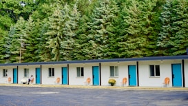 A single-story motel building with a row of five doors and windows, each featuring bright blue doors and orange chairs placed outside. White potted flowers are seen beside a person seated near one of the rooms. The background is dominated by a dense line of tall evergreen trees, adding a natural feel to the setting.