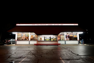 A brightly lit diner with a retro design, featuring a variety of illuminated signs advertising gyros, burgers, hot dogs, and milkshakes. The building has a large overhanging roof with a sign displaying various food options. The surrounding area appears dark, emphasizing the diner’s lighting. A parked car is visible to the left.